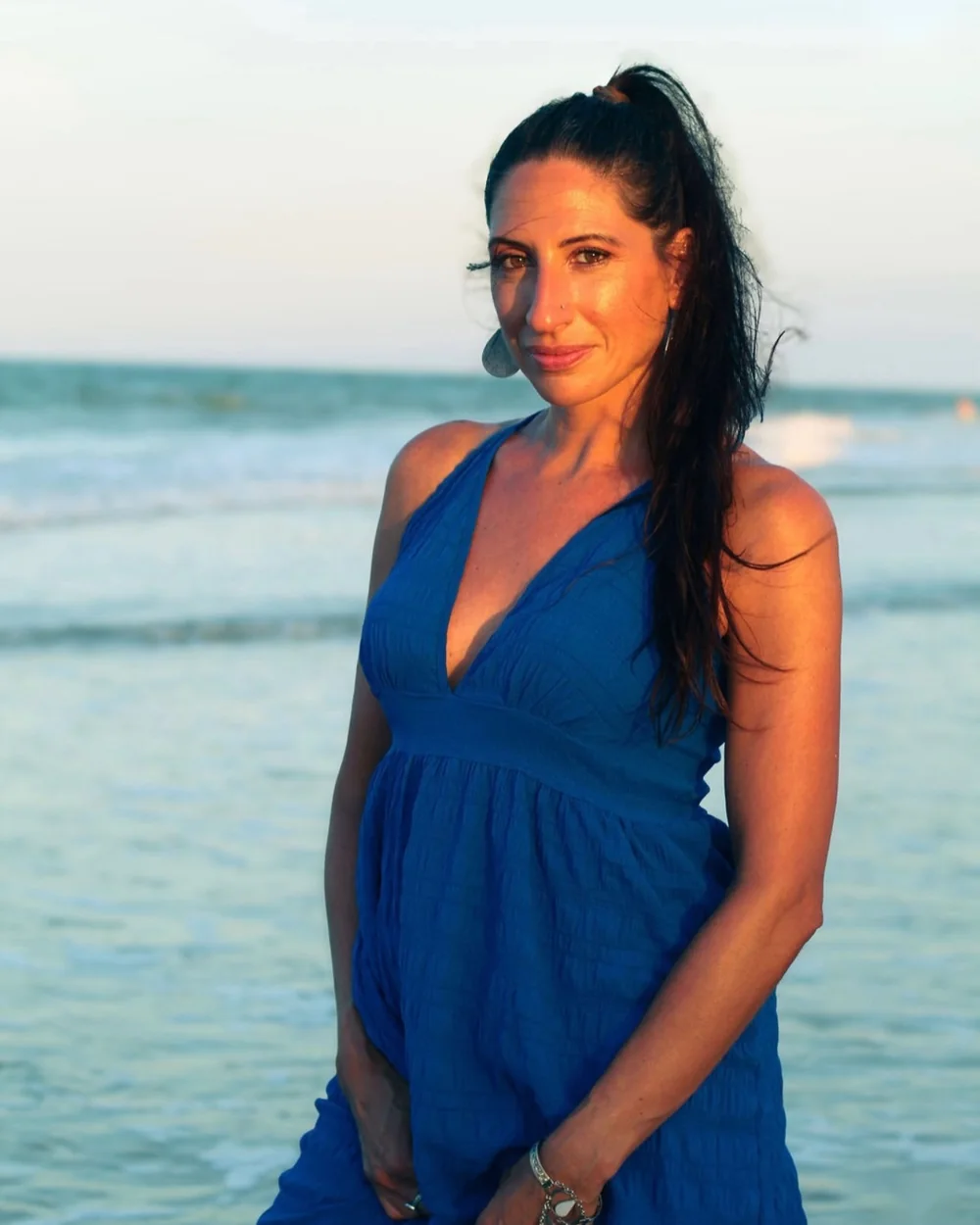 Carla smiling in a blue dress at the water's edge at golden hour