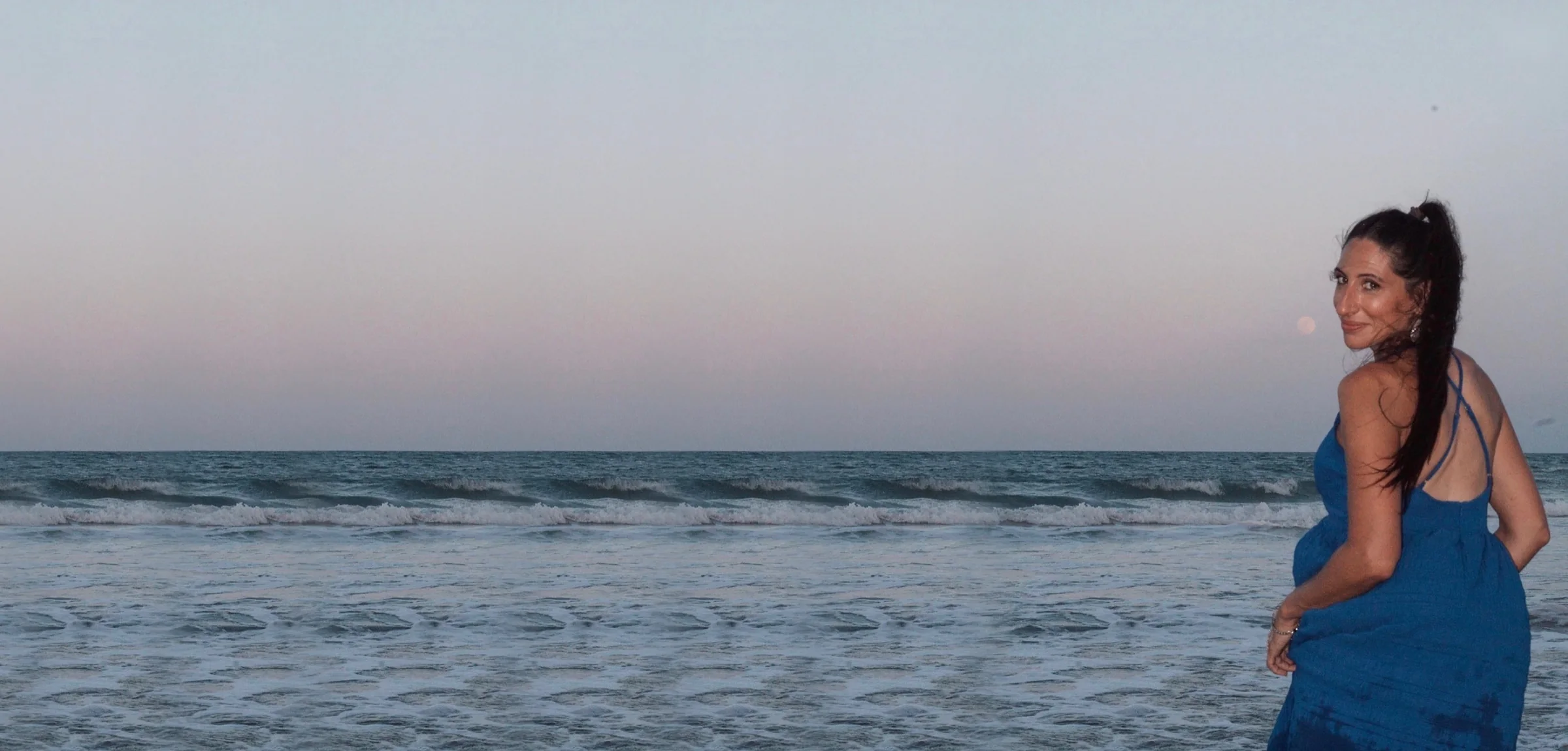 Carla in a blue dress looking back over her shoulder at the ocean, a full moon rising over the water